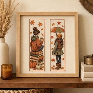 May include: A framed cross-stitch artwork featuring two vertical panels. The left panel shows a person sitting on a stack of books with a mug and pumpkins. The right panel shows a person holding an umbrella with falling autumn leaves.