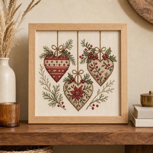 May include: A square framed cross-stitch embroidery featuring three hanging heart-shaped ornaments. The hearts are decorated with red and white patterns, holly leaves, red berries, and a poinsettia flower, all suspended by thin strings against a cream fabric.