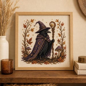 May include: A square framed cross-stitch embroidery featuring a black crow wearing a purple wizard hat and a matching purple cloak with moon symbols. The crow holds a wooden staff, surrounded by autumn leaves and mushrooms on a cream fabric.