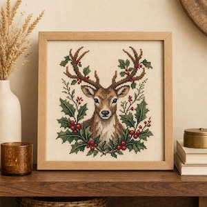 May include: A square cross-stitch embroidery of a deer head centered within a frame of green holly leaves and red berries. The deer has brown fur and antlers, and the design is set against a cream-colored fabric background inside a light wood frame.