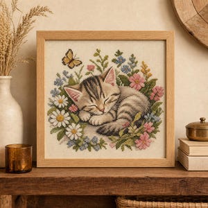 May include: A square cross-stitch embroidery in a light wood frame depicting a sleeping tabby kitten curled up amidst a colourful arrangement of daisies, pink blossoms, and blue flowers, with a small yellow butterfly hovering above.
