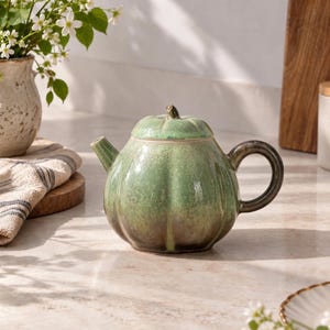 May include: A ceramic teapot shaped like a pumpkin with a glossy green glaze that transitions to a darker brown tone at the base. The teapot features a curved handle, a short spout, and a lid with a small stem-like knob on the top.