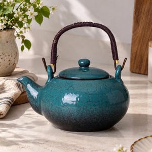 May include: A round ceramic teapot with a deep teal glaze featuring a dark crackle pattern. The teapot has a matching lid, a curved spout, and a dark brown, wrapped handle attached to two metal loops on the top of the vessel's rounded body.
