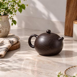 May include: A dark brown, speckled teapot with a rounded body, a curved handle, and a small lid featuring a knob handle. The teapot has a short, simple spout and a textured, matte finish with fine light-colored flecks across the surface.