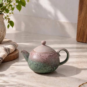 May include: A round ceramic teapot featuring a two-tone glaze with a muted mauve top and a textured sage green base. The teapot includes a matching domed lid with a small knob handle and a curved side handle, showcasing a speckled finish.