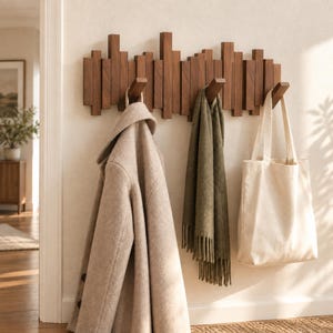 May include: A wall-mounted coat rack made of dark brown wood with vertical slats of varying heights. Three angled hooks extend from the rack, holding a beige wool coat, an olive green fringed scarf, and a plain cream-coloured canvas tote bag.