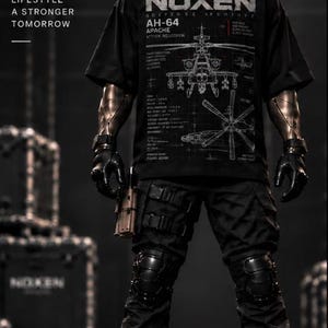 May include: A black t-shirt featuring a white graphic of an AH-64 Apache attack helicopter with technical text, worn with black tactical cargo pants, knee pads, black boots, and a black helmet. The shirt displays the text NOXEN DEFENSE ARCHIVE.