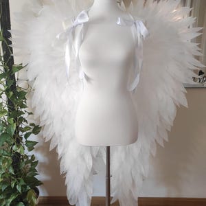 Puede incluir: Grandes alas de ángel blancas expuestas sobre un torso de maniquí blanco. Las alas presentan recortes superpuestos en forma de pluma con una textura ligeramente brillante. Dos cintas de satén blanco están atadas en los hombros.