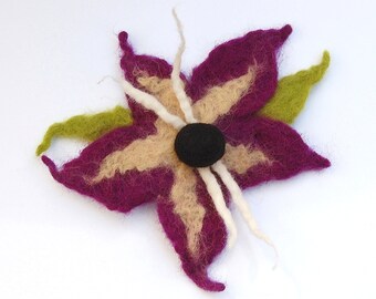 May include: A felt flower brooch with five petals in shades of purple and beige. The centre of the flower is a black felt circle. The flower has white felt stems.