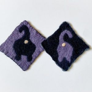 May include: Two felt coasters with a black cat silhouette design. One coaster is purple and the other is a darker shade of purple.