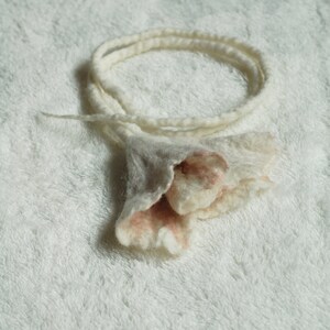 May include: A white and pink felt flower with a long white felt cord. The flower has a ruffled edge and is made of wool.