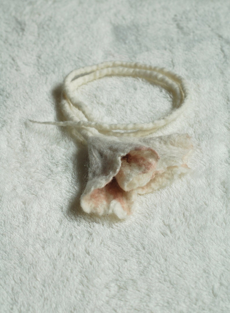 May include: A white and pink felt flower with a long white felt cord. The flower has a ruffled edge and is made of wool.