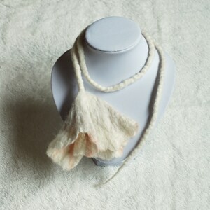 May include: A white and pink felted flower necklace on a white mannequin bust. The necklace is made of wool and has a delicate, feminine look.