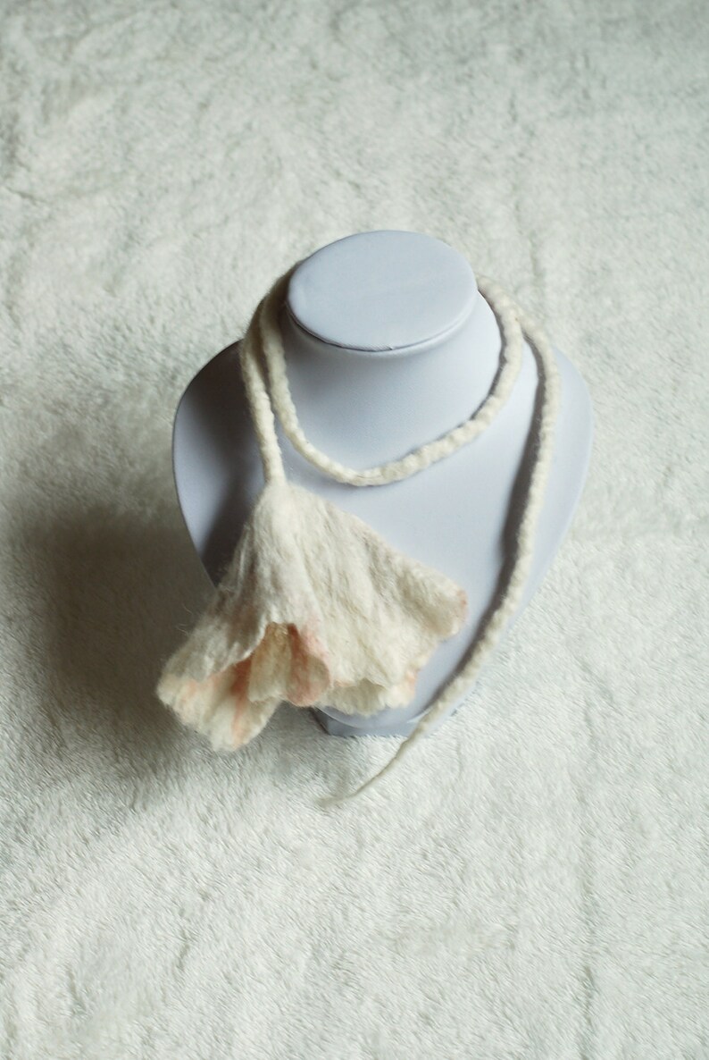 May include: A white and pink felted flower necklace on a white mannequin bust. The necklace is made of wool and has a delicate, feminine look.