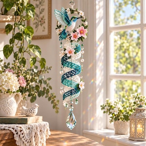 May include: A hanging mobile featuring a crocheted hummingbird in shades of blue and white, perched on a circular wreath of white and pink flowers. Below, a spiral of teal and white lace is accented with white beads and a clear crystal prism.