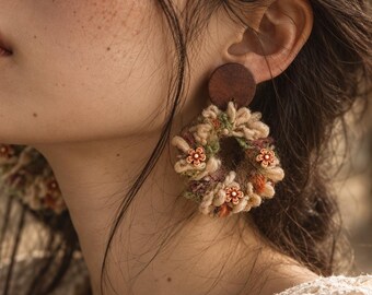 May include: A pair of circular earrings featuring a dark brown wooden stud top connected to a textured, wreath-like hoop made of beige, green, and brown yarn. The hoop is decorated with small, orange, flower-shaped beads with white centres.