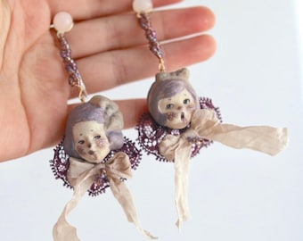 May include: A pair of dangle earrings featuring sculpted doll heads with lavender hair, accented by dark purple lace collars and light beige ribbon bows. Each earring hangs from a single white bead on a gold-toned metal wire.