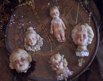 May include: A collection of five necklaces featuring sculpted doll heads and a full doll figure. Each piece is crafted with pale, detailed faces, intricate lace collars, and gold-tone chains. Some pendants include small beads and cross-shaped charms.