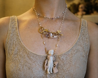May include: A layered necklace featuring a delicate chain with small beads, a gold-toned key charm adorned with amethyst and pearl accents, and a white ceramic cherub figurine pendant draped in light pink fabric, finished with a dangling bead drop.