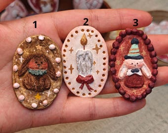 May include: Three oval-shaped clay pins labelled 1, 2, and 3. Pin 1 features a brown rabbit with a green collar. Pin 2 shows a lit candle with a red bow. Pin 3 depicts a rabbit wearing a party hat and bowtie, framed by a dark red beaded border.