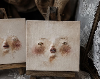 May include: A square canvas painting on a small wooden easel featuring a textured, relief-style face with pale skin, rosy cheeks, green eyes, and parted lips. The artwork uses neutral beige tones and visible brushstrokes to create a dimensional effect.