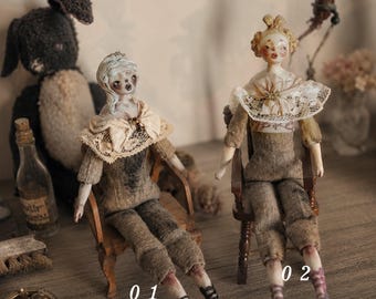 May include: Two handcrafted dolls seated on small wooden chairs. Doll 01 has pale skin, light hair, and wears a beige lace-trimmed top with brown trousers. Doll 02 has blonde hair, a lace collar, and wears a beige top with brown trousers and lace-up shoes.