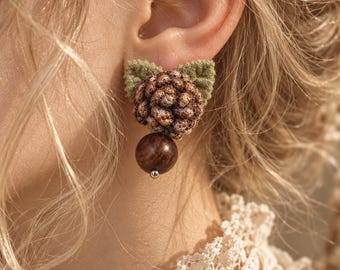 May include: A stud earring featuring a textured, brown, spherical pinecone-like element nestled against two small, sage green fabric leaves, with a smooth, dark brown wooden bead dangling directly beneath the pinecone component.