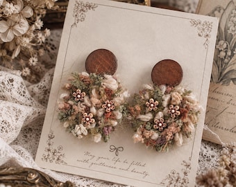 May include: A pair of earrings featuring dark brown circular wooden studs attached to textured, circular fibre clusters in shades of green, cream, and pink, accented with small wooden flower charms, displayed on a card with the text May your life be filled with gentleness and beauty.