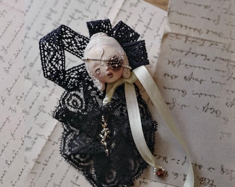 May include: A brooch featuring a pale, sculpted doll-like face with a dark floral eye patch, adorned with black lace ruffles, a cream-coloured ribbon, and small beaded accents. The piece is constructed with intricate fabric layers and a central figure.