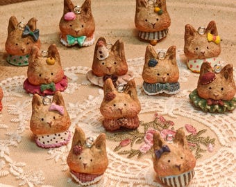 May include: A collection of twelve small, brown, sculpted animal charms, each featuring a unique headpiece or collar. The charms are shaped like cat or rabbit heads with painted facial features and metal loops at the top for hanging.