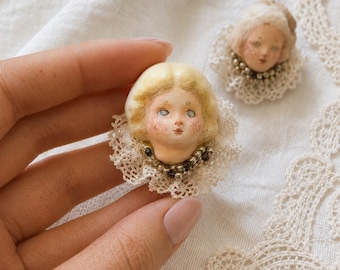 May include: Two small doll head brooches featuring pale faces with rosy cheeks, blonde hair, and blue eyes. Each head is framed by a circular white lace collar and accented with a necklace of small, dark metallic beads around the neck area.