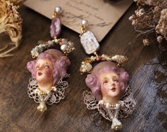 May include: A pair of dangle earrings featuring sculpted faces with mauve hair, accented by white lace collars, small pearls, and clear beads. Each earring hangs from a gold-tone post with a single pearl and a rectangular mauve stone.