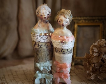 May include: Two handcrafted fabric dolls featuring sculpted faces and intricate hairstyles. The left doll wears a lace-trimmed bodice with light blue bows, while the right doll features a peach-coloured bodice with matching bows and lace accents.