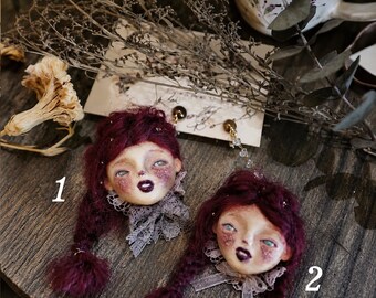 May include: Two handmade doll head brooches featuring deep burgundy hair styled in braids with small pom-poms. Each doll has a pale face, dark lips, and a grey lace collar. The brooches are attached to a white card with handwritten text.