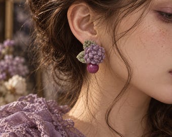 May include: A crochet earring featuring a cluster of purple yarn flowers with two small green leaves and a single smooth, dark magenta spherical bead dangling from the bottom, attached to a post backing for secure placement on the earlobe.
