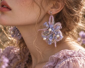 May include: A pair of statement earrings featuring intricate lavender and soft green yarn loops, textured fibre clusters, and small white faux pearls. The earrings are attached to silver-toned metal posts and exhibit a layered, multi-dimensional design.