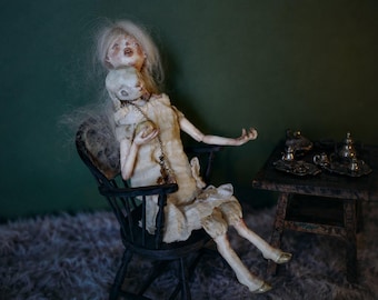 May include: A pale, articulated doll with long white hair sits in a black wooden chair, wearing a cream-coloured dress. The doll holds a small, white, rabbit-like head on a chain. Beside the chair is a small, dark wooden table with miniature silver-toned tea service items arranged on top.