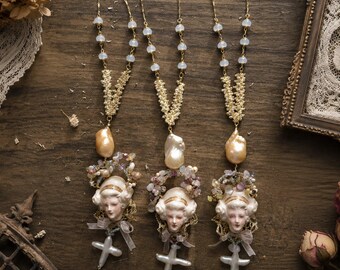 May include: Three gold-tone necklaces featuring chains with small white beads, a central gold-tone textured V-shape, a large irregular cream-coloured pearl, a cluster of small multicoloured gemstones, a ceramic face pendant, a small ribbon, and a cross-shaped pearl.