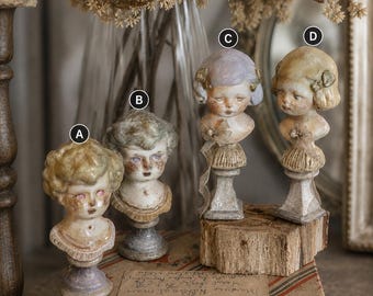 May include: Four small decorative busts labelled A, B, C, and D, each featuring a sculpted child's head with distinct hair colours and facial expressions, mounted on individual pedestals with a weathered, aged finish and small embellishments.