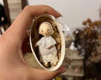 May include: A small doll with a pale face and light hair, wearing a white lace collar and a textured beige dress, nestled inside a small oval wooden box lined with brown paper. The doll has delicate painted features and gold-toned shoes.