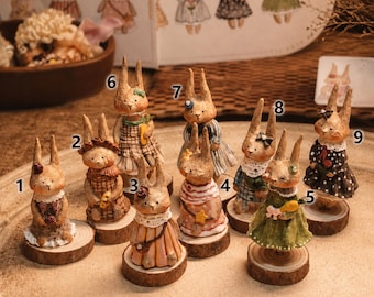May include: A collection of nine small rabbit figurines, each numbered 1 through 9, standing on individual circular wood slices. The rabbits are dressed in various outfits, including dresses and shirts with patterns, and are displayed on a light surface.
