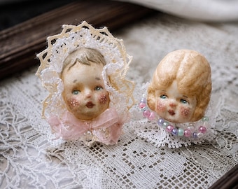 May include: Two decorative doll heads featuring painted facial features with rosy cheeks. One head is framed by a white lace bonnet with a pink ribbon bow. The other head is adorned with a collar of small, multicoloured iridescent beads.