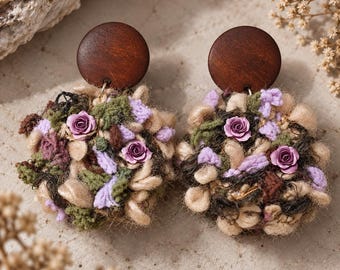 May include: A pair of earrings featuring circular dark brown wooden studs attached to round, textured dangles. The dangles are composed of mixed yarn fibres in shades of olive green, beige, and lavender, accented with small, light purple rose embellishments.