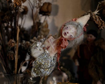 May include: A handcrafted doll with a pale, textured face, grey hair, and a pointed cap tied with a thin red ribbon. The doll wears a bodice adorned with intricate silver and gold embroidery and small beads, featuring a delicate, weathered appearance.