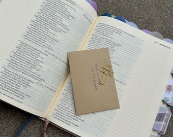 May include: An open Bible with colorful page tabs and a small, rectangular, light brown paper envelope resting on the pages. A gold-colored metal cross-shaped paperclip with a heart cutout is attached to the envelope, which has the text "Read me when you are scared" printed on it.