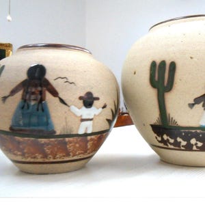May include: Two round, cream-colored ceramic vases featuring hand-painted desert scenes. Each vase depicts figures in blue and white clothing, green cacti, and brown ground details. The base of each vase has a dark brown band with a repeating pattern.