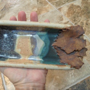 May include: Handmade ceramic rectangular dish with a beige rim and interior with blue, brown, and tan hues. One end features a braided design, while the other has textured brown leaf accents. A unique decorative piece.