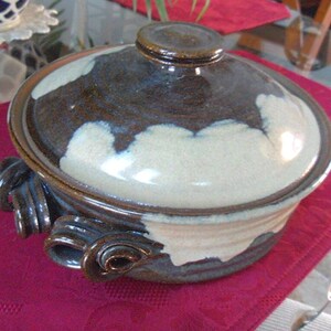 May include: A ceramic casserole dish with a lid. The dish is primarily brown with white and cream-colored accents. It has decorative handles on the sides. The lid has a small knob on top. The dish is on a red tablecloth.