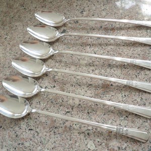 May include: Six silver spoons arranged diagonally. Each spoon has a rounded bowl and a long, slender handle with a decorative floral design near the end. The spoons are reflective, and the background is a speckled, light brown surface.