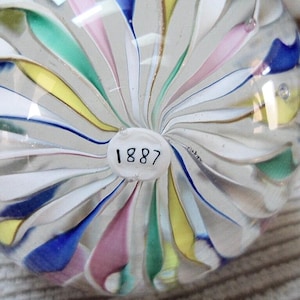 May include: A clear glass decorative object with a swirl design. The object features white, blue, pink, yellow, and green colors. The number "1887" is printed on a white circular label on the bottom of the object.